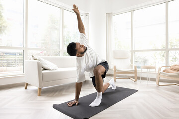 Serious young Black man doing morning exercises at home, keeping twisting yoga asana on mat, caring...
