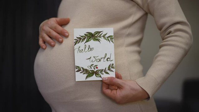 Slow motion. Close-up. Profile of an unrecognizable pregnant woman holding a birthday card and rubbing her belly with hand, while standing indoors