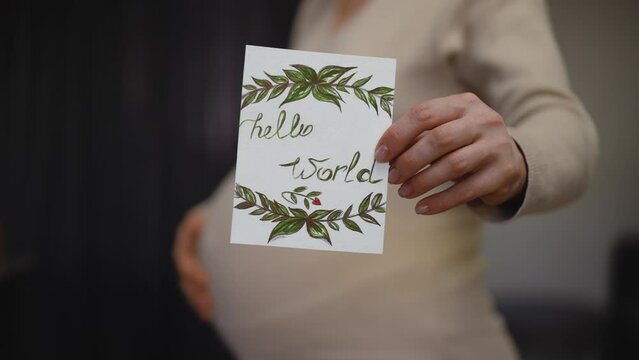 Close-up. Slow motion. Hello world. Profile of an unrecognizable pregnant woman, holding a birthday card with one hand and holding it out towards the camera, while the other hand rubs her belly, while