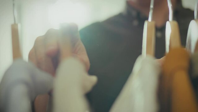 A man, standing in front of a rack with wooden hangers, carefully examines the clothes. His hands, close-up, carefully move the hangers. Reflects the modern concept of men's fashion