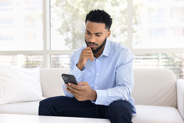 Serious thoughtful young African man using financial service, Internet application on mobile phone, typing message on smartphone, touching chin, sitting on couch at home, reading