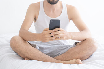 A bearded man with his face hidden, chatting on his smartphone, sitting on bed in lotus pose....