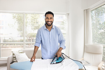 Cheerful attractive young African American homeowner man ironing linen on board at home, enjoying...