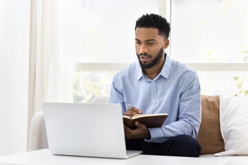 Serious African student man watching online lecture, training seminar on laptop at home, writing notes, summary of webinar, studying on Internet, doing learning homework, speaking on video call