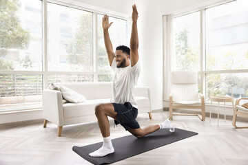 Happy positive sporty African guy training body on yoga mat, doing lunges with raised hands,...