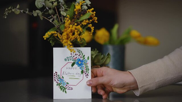 Slow motion. Close-up. A woman's hand places a handmade Mother's Day card near a bouquet of spring flowers in a glass vase standing on a table indoors