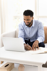 Cheerful young Black student guy watching online training lecture on laptop, writing notes,...
