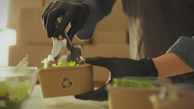 A warehouse worker packs fresh salads into disposable containers in an industrial kitchen. Food delivery employee packing food order into paper container sustainable business idea without waste