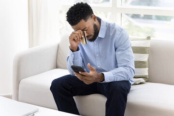 Sad tired young African bank client holding mobile phone and credit card, finding financial problems, lack of money, bad ecommerce service, fraud, thinking on overspending, bankruptcy