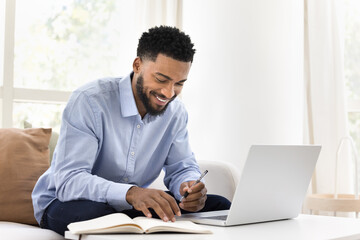 Positive young African online student man studying on Internet, sitting at laptop at home, writing notes, watching webinar, lecture, training class, doing learning homework, smiling