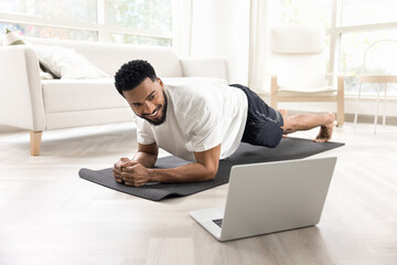 Cheerful strong athletic African man keeping static plank on yoga mat at laptop, looking at screen,...