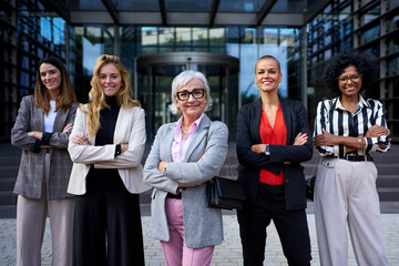 Group of successful diverse race and ages business women entrepreneurs standing in formal wear with arms crossed looking at camera. Happy and empowered adult female professionals co-workers outdoors - obrazy, fototapety, plakaty