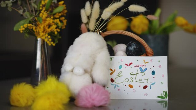 Close-up. For the Easter celebration, there is a glass vase with spring flowers, an Easter bunny, a basket of Easter eggs and a handmade Easter card on the table