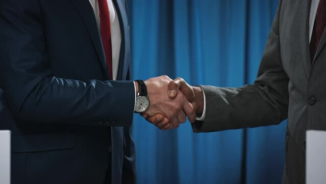 Medium close-up shot of midsection and hands of anonymous diverse male politicians in formal suits and ties standing on stage at tribunes, shaking hands in front flashing cameras after election debate