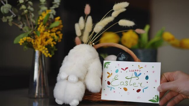 Close-up. Celebration of Easter. On the table in the apartment there is a glass vase with spring flowers, an Easter bunny, and a basket with Easter eggs. A woman's hand places a handmade Easter card