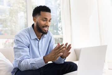 Fototapeta premium Positive handsome young African business man talking on work video call on laptop, speaking at computer, moving hands, convincing client on online conference meeting