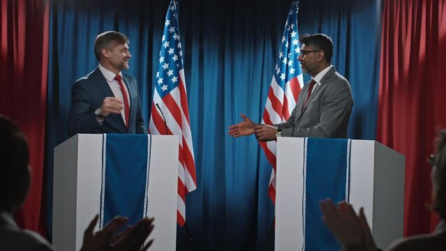 Full shot of Caucasian and Indian American presidential candidates finishing debates, thanking each other, shaking hands, audience giving standing ovation, and photographer taking photos with flash