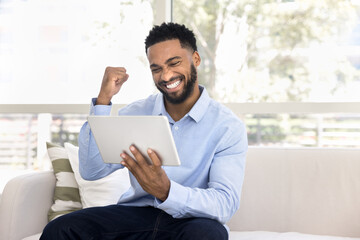 Cheerful overjoyed young African woman celebrating triumph, success, win, looking at tablet, making hand yes gesture, smiling, laughing, getting happy, excited, enjoying home Internet communication