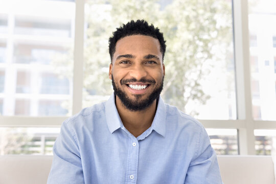 Cheerful attractive African freelance business man talking on video conference call from home, sitting on couch, looking at camera with toothy smile. Screen view head shot portrait