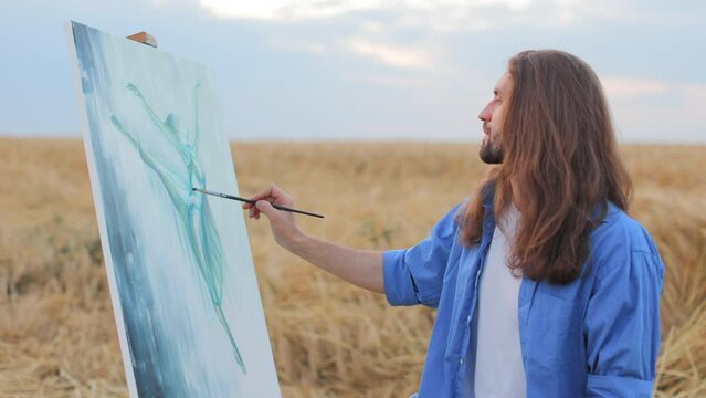 Man with long dark hair holding brush and painting dancing woman on canvas while standing in field of rye. Male artist creating masterpiece while getting inspired by nature and female beauty.