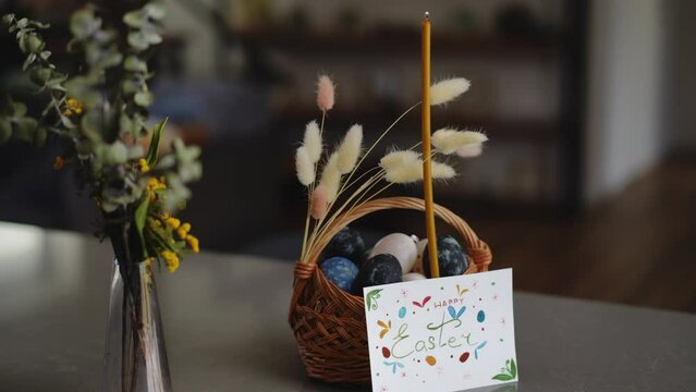Close-up. Happy Easter holiday. A burning wax candle stands in a decorated basket with Easter eggs near a glass vase with spring flowers. Near the basket standing nearby a handmade Easter card