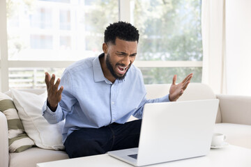 Fototapeta premium Angry concerned young African business man shouting at laptop, getting bad news, problems with work software, service wrong work, losing out, feeling stressed, frustrated, nervous