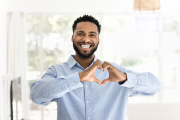 Positive attractive young African man making hand heart gesture, joining fingers at chest,...