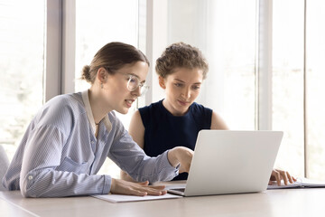 Two young office employees working together use laptop, workmate helps to apprentice with corporate...