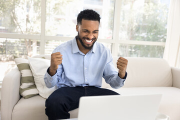 Happy excited young African business man making hand yes gesture at laptop, celebrating triumph, good news, success, achievement, enjoying communication, Internet technology