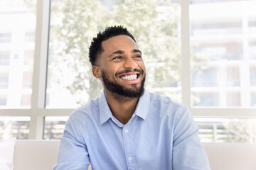 Cheerful attractive young Black man in pale blue shirt looking away with perfect toothy smile in deep positive thoughts, laughing, showing white teeth, promoting dental care