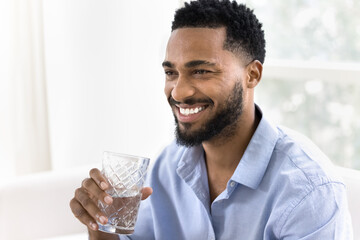 Happy attractive young African man drinking clear pure water, holding glass with cold beverage, looking away with toothy smile, enjoying healthy aqua balance, hydration, refreshing