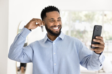 Happy new African American realtor holding key, taking self video for blog n smartphone, promoting...