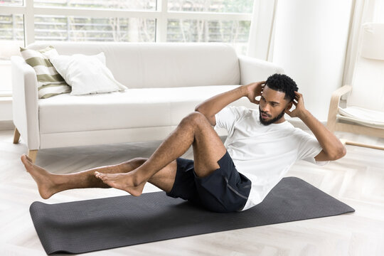 Focused young African athlete guy twisting body in boat pose during morning routine fitness activity, doing abs workout, training core muscles at home, exercising on mat