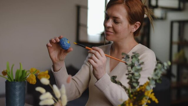 A pregnant woman holds an Easter egg in her hand and touches up the ornament on it with a paintbrush. The woman nods her head smiling