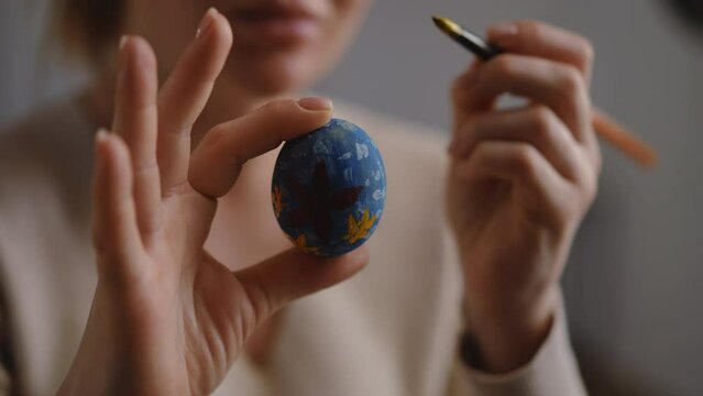 Close-up. A woman holds an Easter egg in her hand and touches up the ornament on it with a paintbrush
