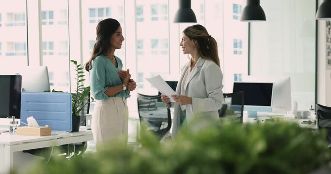 Businesswomen discuss details of task and collaborative project standing in co-working office. Two women colleagues lead business conversation in corporate workspace. Workday, workflow, communication