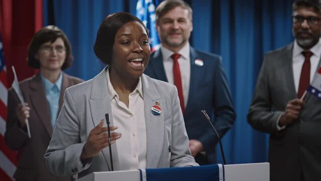 Medium shot of enthusiastic young African American female presidential candidate standing at rostrum, delivering energetic political manifesto for US elections, diverse team with flags in background