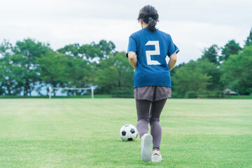 グランドでサッカーボールをドリブルするユニフォームを着た女性の後ろ姿
