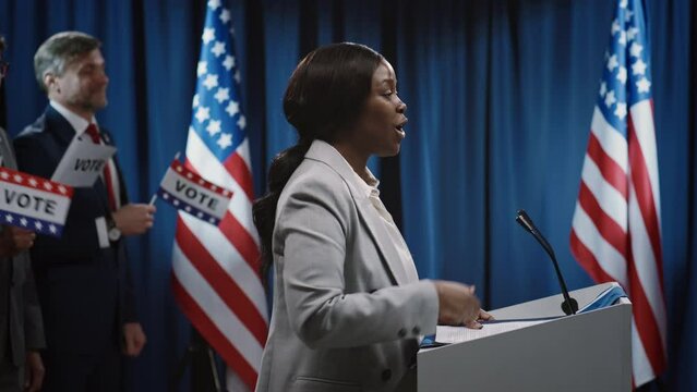 Medium side footage of young African American female candidate for president or mayor from Democratic party standing at tribune and delivering energetic policy speech to public in hall