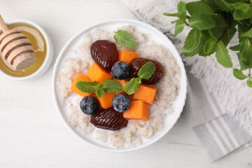Delicious barley porridge with blueberries, pumpkin, dates and mint served with honey on white table, flat lay