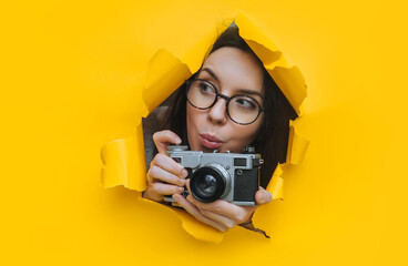 A paparazzi girl in glasses with a rare SLR camera looks out from her hiding place and carefully looks at what is happening. Yellow paper, torn hole. Tabloid press.Looking for a story for stock photos - obrazy, fototapety, plakaty