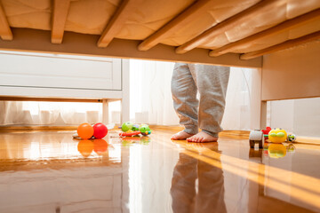 Baby standing near a crib with toys on the floor. View from under the bed