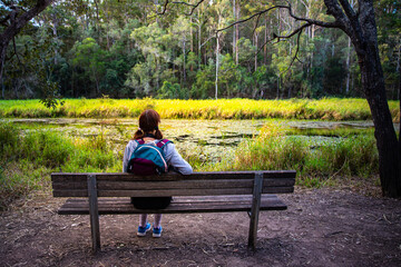 pretty girl with a backpack resing on a bench enjoys the view of enoggera reservoir in brisbane, queensland, australia; warm sunset in a park	