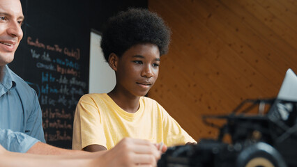 African boy using laptop to program code or prompt with car model placed while teacher and...