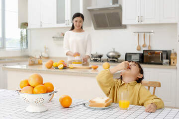 Asian daughter drinks freshly squeezed orange juice with bread, mother is making freshly squeezed orange juice