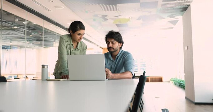 In modern office, Indian colleagues collaborate, working use laptop. Serious workmates, mentor and apprentice discuss project details, share ideas, provide helpful information, learn new corporate app
