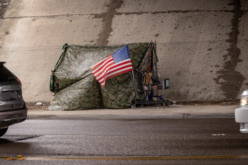 An American flag at a homeless tent made of camouflage tarp at a road underpass with a car visible