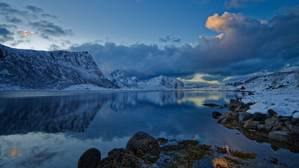 Old red cabin on snow covered rocky terrain by the lake surrounded by the snowy mountains. Lofoten Islands, Northern Norway.