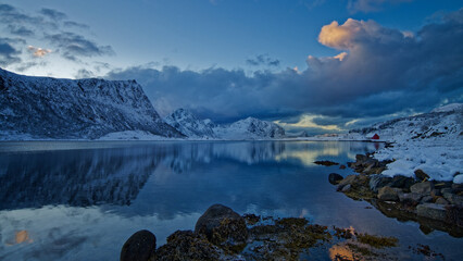 Old red cabin on snow covered rocky terrain by the lake surrounded by the snowy mountains. Lofoten...