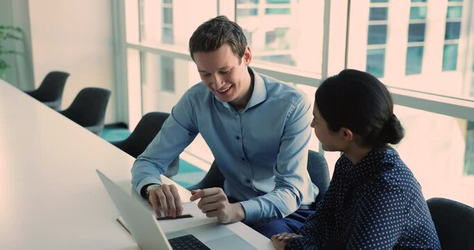 Young man share marketing strategy, working on collaborative project sit at desk with laptop engaged in teamwork with young Indian female workmate. Sales manager explain offer to client met in office